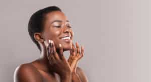 Happy woman with closed eyes touching soft smooth skin on her cheeks over grey background